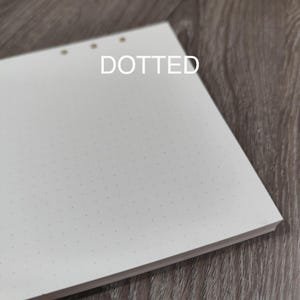 May include: A stack of white, dotted paper with three holes at the top. The word "DOTTED" is printed in white across the top of the paper. The paper is on a wooden surface.