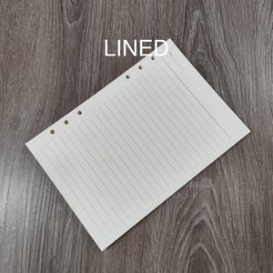 May include: A cream-coloured, lined paper sheet with three holes for binding. The word "LINED" is printed in white at the top. The paper is angled on a wooden surface.
