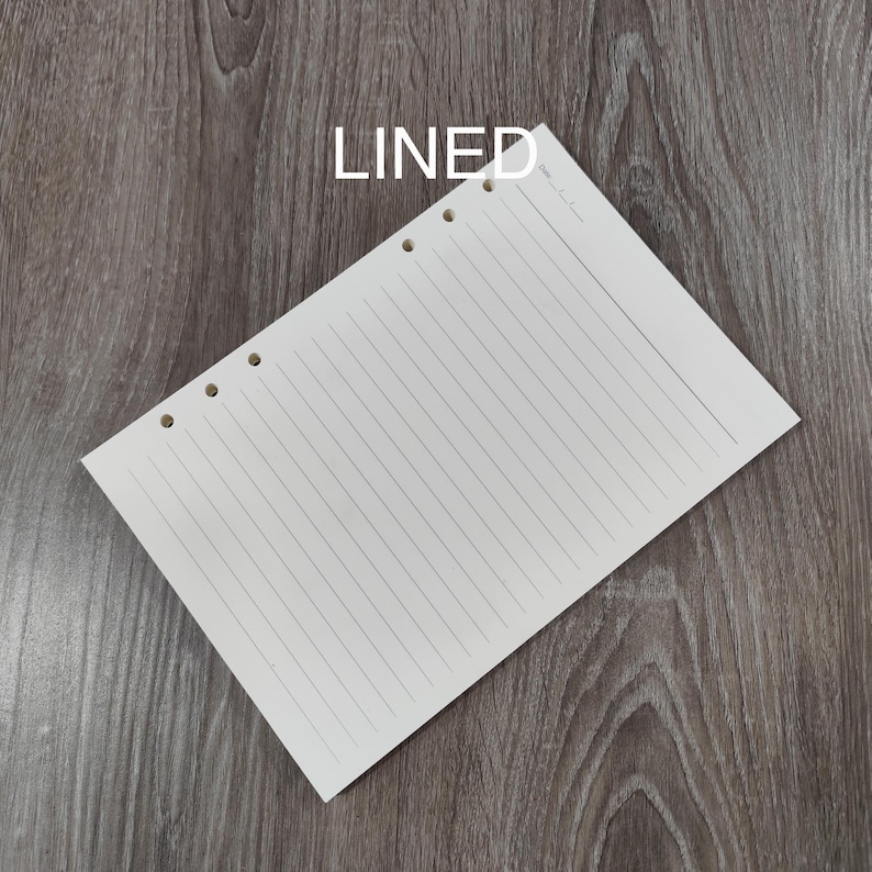 May include: A cream-coloured, lined paper sheet with three holes for binding. The word "LINED" is printed in white at the top. The paper is angled on a wooden surface.