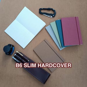 May include: A collection of B6 slim hardcover notebooks in various colors including maroon, teal, sage green, light gray, and brown. The set includes an open white notebook, a black carabiner, a dark brown pen case with pens, and an ink bottle.
