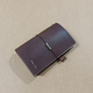 May include: A dark brown leather notebook cover featuring a vertical black elastic closure band with a metallic brass-toned bead. The front cover is embossed with the name Pham Duy in a simple serif font, and a small leather pen loop is attached.
