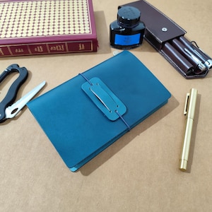 May include: A teal leather journal with a matching leather strap closure, positioned alongside a brass fountain pen, a black ink bottle with a blue label, a brown leather pen case, a folding pocket knife, and a red hardcover book.