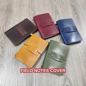 May include: Five leather field notes covers in various colors: brown, burgundy, blue, yellow, and olive green. Each cover has a strap closure and a brass-colored metal accent. The text "FIELD NOTES COVER" is displayed at the bottom.