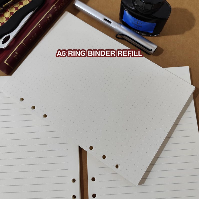 May include: White A5 ring binder refill paper with a dotted grid pattern. The paper has pre-punched holes for a binder. The text "A5 RING BINDER REFILL" is printed in red. A pen and ink bottle are visible in the background.