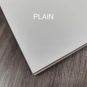 May include: A white, rectangular object with the word "PLAIN" printed in the centre. The object is angled, showing its edge, and is set against a wooden surface.