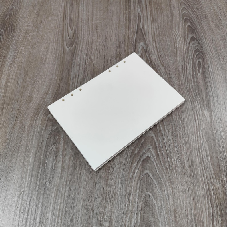 May include: A stack of white, blank paper with three holes punched along the left edge. The paper is rectangular and appears to be a notepad or a stack of loose-leaf paper. The paper is on a wooden surface.