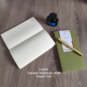 Könnte beinhalten: Ein 2er-Pack Traveler Notizbuch-Nachfüllungen, reguläre Größe, mit leeren Seiten. Ein grünes Notizbuch, ein goldener Stift und eine Flasche schwarzer Tinte sind ebenfalls zu sehen. Der Text lautet: "2-pack Traveler Notebook Refill Regular Size."