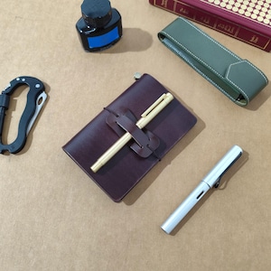 May include: A dark brown leather notebook with a brass pen held by a leather strap, a black ink bottle, a green leather pen case, a silver fountain pen, and a black carabiner. The items are arranged together, showcasing stationery and utility tools.
