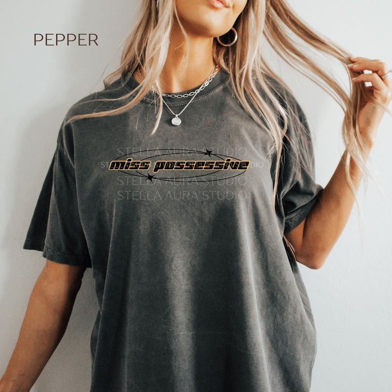 May include: A gray oversized t-shirt with the text 'miss possessive' in a black oval with gold stars around it.