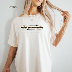 May include: Ivory-colored t-shirt with the text "miss possessive" in a black and yellow oval design. The shirt also has the text "STELLA AURA STUDIO" below the oval. The shirt is a short-sleeved, casual style.