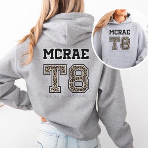 May include: A gray hoodie with the name "MCRAE" in black block letters above the number "T8". The "T8" is in a leopard print design. The words "STELLA AURA STUDIO" are printed below the number.