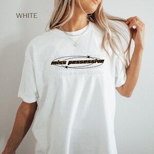 May include: White t-shirt with black text that reads 'miss possessive' in a circle with black stars around it.