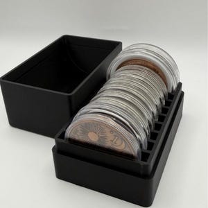 May include: A black rectangular storage box with a removable lid, containing a row of circular coins encased in clear protective plastic capsules. The visible coin features a sunburst design and the number 1, with a metallic copper-toned finish.