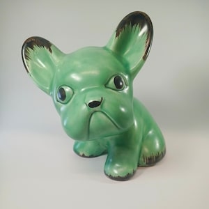 May include: A ceramic French Bulldog figurine, seated and in a light green glaze. The ears, paws, and nose have dark brown detailing. The dog has black eyes and a slightly grumpy expression. A decorative item.