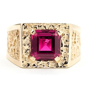 Puede incluir: Anillo dorado con una gran gema cuadrada de color magenta en el centro. El anillo tiene una banda y un engaste texturizados, creando una estética vintage o antigua. La gema es el punto focal, captando la luz.