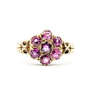May include: A gold ring featuring a cluster of round, deep pink gemstones arranged in a floral pattern. The ring's band has ornate, filigree-style detailing on the sides, and the gemstones are set in a gold setting.