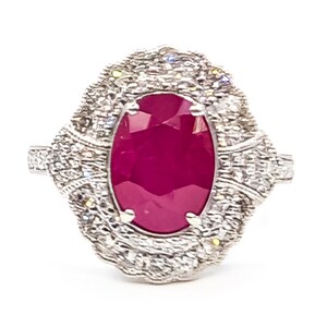May include: A ring featuring a large, oval-cut, deep red ruby set in the center. The ruby is surrounded by a textured, scalloped halo of small, clear, sparkling stones set in a silver-toned metal band with intricate detailing on the sides.