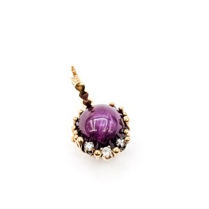 May include: A gold-colored pendant featuring a large, round, deep purple gemstone. The gemstone is surrounded by a gold setting with small, clear accent stones. The pendant has a decorative loop for a chain.