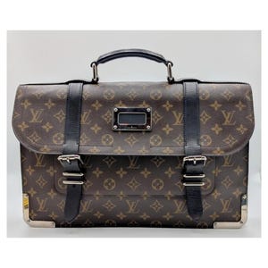 May include: A brown briefcase featuring the iconic monogram pattern in dark brown and tan. The bag includes a top handle, two black leather straps with silver-tone metal buckles, a central silver-tone rectangular plate, and silver-tone corner protectors.