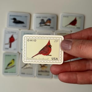 May include: A collection of rectangular, postage stamp-style magnets featuring various bird illustrations. The central magnet showcases a red Northern Cardinal bird with the text "OHIO" and "USA". The stamps have a cream background and a scalloped border.