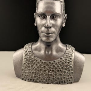Könnte beinhalten: Silberfarbene Büstenskulptur einer Person mit kurzen Haaren und einem detaillierten Oberteil. Die Skulptur hat einen ernsten Ausdruck und ist vor einem dunklen Hintergrund platziert. Das Oberteil weist ein Muster aus verbundenen Kreisen auf.