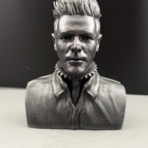 Könnte beinhalten: Silberfarbene Büstenskulptur einer Person mit kurzen, stacheligen Haaren und einem Hemd mit Kragen. Die Skulptur hat einen Stachelkragen und einen neutralen Gesichtsausdruck. Die Büste ist vor einem dunklen Hintergrund platziert.