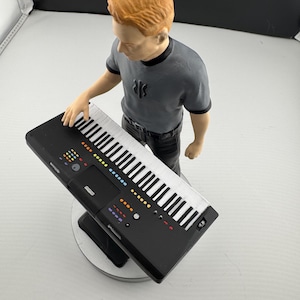 Könnte beinhalten: Figur einer Person mit orangefarbenem Haar, die ein graues T-Shirt und eine schwarze Hose trägt und über einer schwarzen Tastatur mit weißen Tasten steht. Die Tastatur hat bunte Knöpfe und Lichter. Die Figur steht auf einem silbernen Sockel.