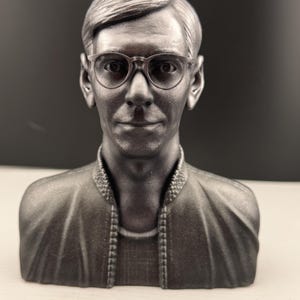 Könnte beinhalten: Silberfarbene Büste einer Person mit kurzen, zurückgekämmten Haaren und einer Brille mit klarem Rahmen. Die Büste zeigt eine Jacke mit detailliertem Kragen und eine zarte Halskette. Der Hintergrund ist dunkel.