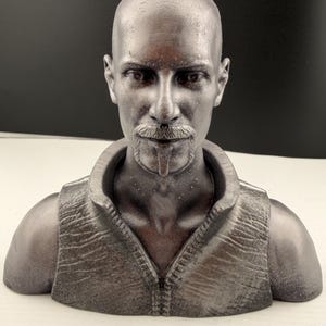 Könnte beinhalten: Silberfarbene Büste eines Mannes mit rasiertem Kopf, Schnurrbart und Spitzbart. Die Skulptur zeigt den Mann von der Brust aufwärts, mit einer Weste mit Reißverschluss und einem Hemd mit Kragen. Die Büste steht vor einem neutralen Hintergrund.