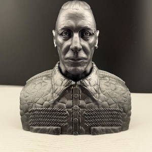 Könnte beinhalten: Metallgraue Büstenskulptur eines Mannes mit rasiertem Kopf und Piercing. Die Skulptur zeigt eine detaillierte Jacke mit strukturiertem Muster und Knöpfen. Die Büste steht vor einem dunklen Hintergrund.