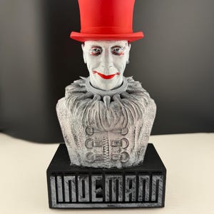 Könnte beinhalten: Eine Büste einer Figur mit weißer Gesichtsbemalung, roten Lippen und einem roten Zylinder. Die Figur hat einen Rüschenkragen und eine Jacke mit dekorativen Knöpfen. Die Büste ist auf einem schwarzen Sockel mit dem Namen "INDEMANN" in Blockbuchstaben montiert.