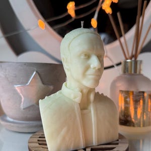 Könnte beinhalten: Eine cremefarbene Kerze in Form einer Büste einer Person, die auf einem Holzuntersetzer steht. Die Kerze hat einen Docht und befindet sich vor einem dekorativen Hintergrund, einschließlich eines sternförmigen Behälters und eines Diffusors.