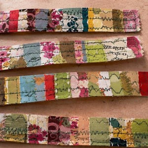 May include: Four horizontal strips of fabric featuring a patchwork design of various floral, solid, and patterned segments in shades of red, pink, yellow, green, and blue, all joined together with visible dark green zigzag machine stitching.