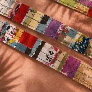 May include: Two strips of fabric featuring a patchwork design with various colours including red, yellow, blue, purple, and black. The fabric is decorated with a repeating pattern of small dark green stitched lines running horizontally across the surface.