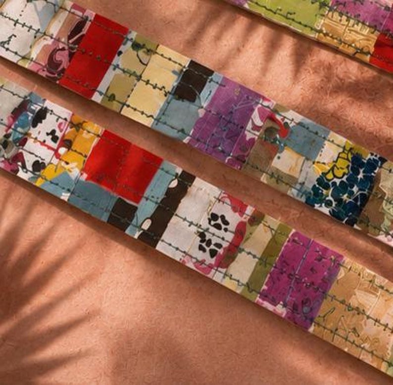 May include: Two strips of fabric featuring a patchwork design with various colours including red, yellow, blue, purple, and black. The fabric is decorated with a repeating pattern of small dark green stitched lines running horizontally across the surface.
