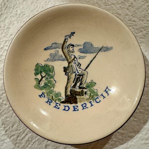 May include: A round, off-white ceramic dish featuring a hand-painted illustration of a soldier standing with a rifle and a cannon. The scene includes green trees and blue clouds, with the word FREDERICIA printed in blue capital letters below.