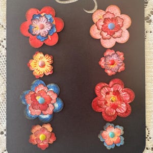 May include: A black card with a decorative swirl at the top, featuring several colorful paper flower embellishments. The flowers are in shades of red, pink, yellow, and blue, with varying sizes and center details.