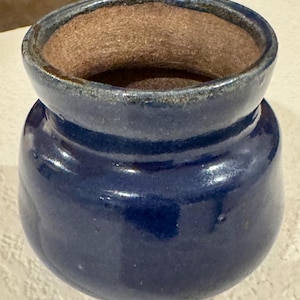 May include: A small, rounded ceramic pot with a deep, glossy cobalt blue glaze on the exterior. The vessel features a wide, thick rim that transitions into a textured, unglazed brown interior surface, showcasing a contrast between the smooth blue finish.