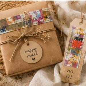 May include: A square gift box wrapped in brown paper with a colourful patterned strip, tied with twine and a round tag reading happy mail with a heart. Beside it is a rectangular tag with a colourful strip and the text no. 01, tied with twine.