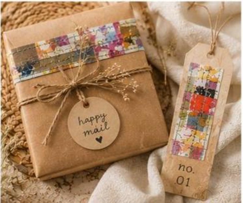 May include: A square gift box wrapped in brown paper with a colourful patterned strip, tied with twine and a round tag reading happy mail with a heart. Beside it is a rectangular tag with a colourful strip and the text no. 01, tied with twine.