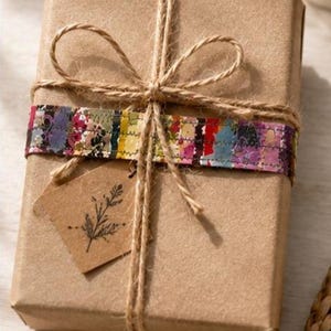 May include: A rectangular gift box wrapped in plain brown paper, secured with natural jute twine tied in a bow. A horizontal strip of multicoloured floral fabric is wrapped around the centre, and a small brown paper tag with a black leaf illustration.