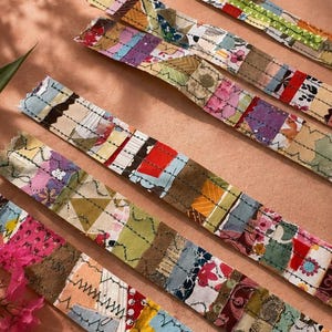 May include: Several long fabric strips featuring a patchwork of various colourful textile scraps in shades of red, pink, yellow, green, and blue. Each strip is reinforced with visible dark thread stitching in straight and zigzag patterns.