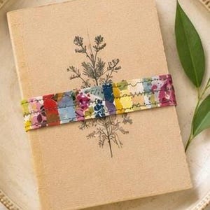 May include: A rectangular tan notebook with a black botanical line drawing of a plant on the cover. A horizontal fabric band featuring a multicoloured floral and abstract pattern is wrapped around the centre of the notebook, secured with visible stitching.