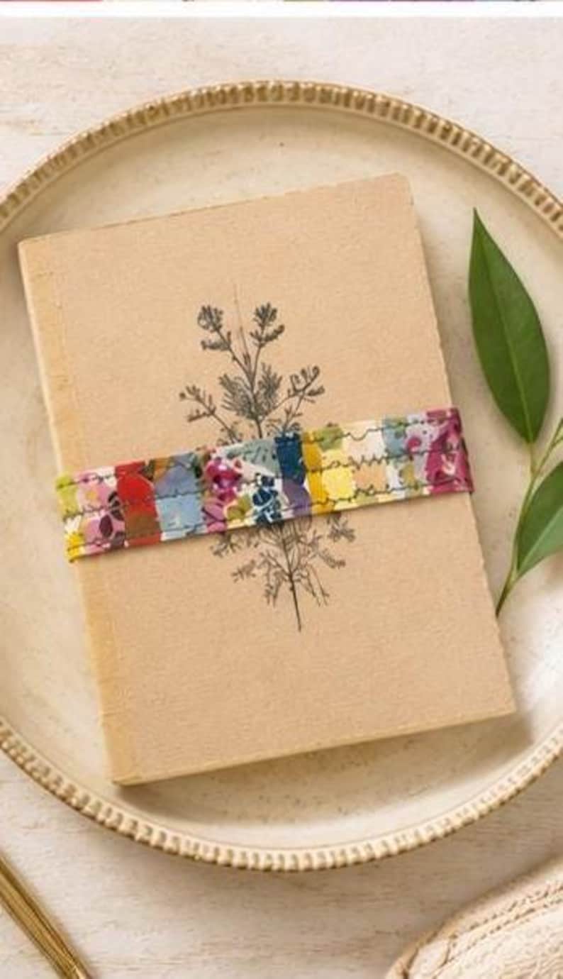 May include: A rectangular tan notebook with a black botanical line drawing of a plant on the cover. A horizontal fabric band featuring a multicoloured floral and abstract pattern is wrapped around the centre of the notebook, secured with visible stitching.