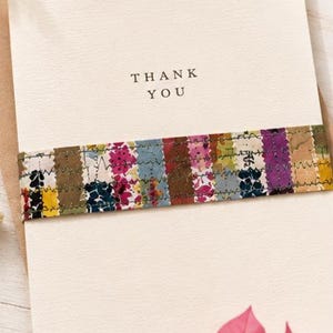 May include: A cream-coloured thank you card featuring a horizontal band of multicoloured fabric strips with a floral pattern. The fabric band is secured with visible zigzag stitching. The words THANK YOU are printed in black ink on the card.
