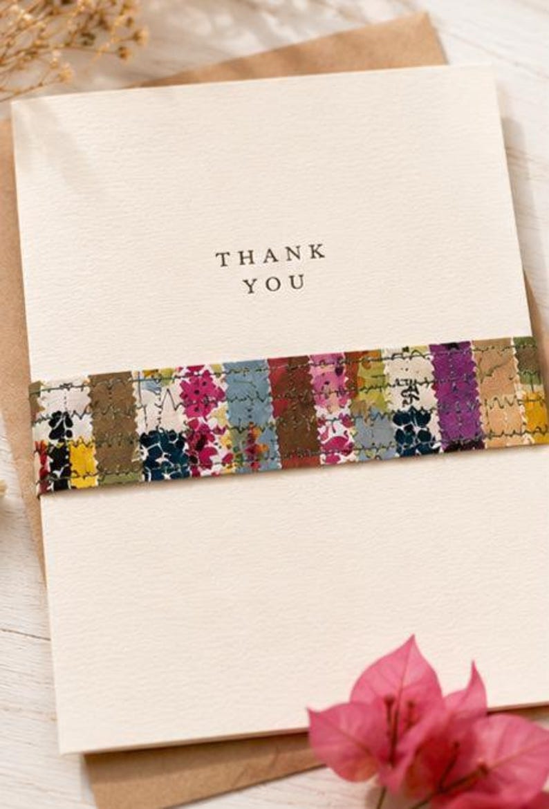 May include: A cream-coloured thank you card featuring a horizontal band of multicoloured fabric strips with a floral pattern. The fabric band is secured with visible zigzag stitching. The words THANK YOU are printed in black ink on the card.