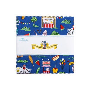 May include: A stack of square fabric pieces featuring a blue background with a carnival-themed print including hot dogs, corn dogs, watermelon slices, and fair booths. A white paper band wraps the center with the text Riley Blake Designs and Wisconsin Best In Show with a trophy icon.