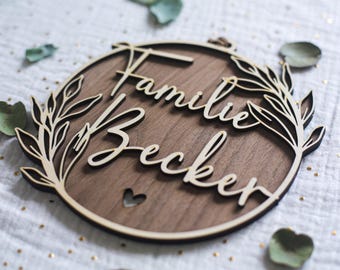 May include: A circular wooden wall sign featuring a dark brown wood grain background with light-colored, laser-cut lettering that reads "Familie Becker." The design includes a decorative leaf border on the sides and a small heart cutout below.