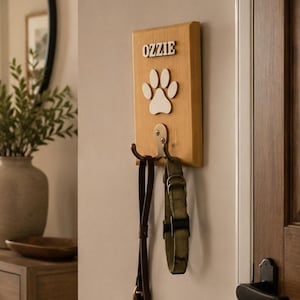 May include: A rectangular light wood wall-mounted hook featuring the name OZZIE in raised letters above a white paw print cutout. A metal hook is attached to the bottom, holding a dark brown leather lead and a green fabric pet collar.