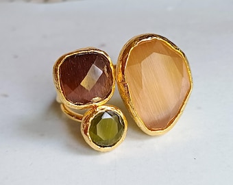 May include: Three gold rings featuring large gemstones in brown, olive green, and peach. The rings have textured gold bands. The gemstones are a square brown, a round olive green, and a large, irregular peach-colored stone.